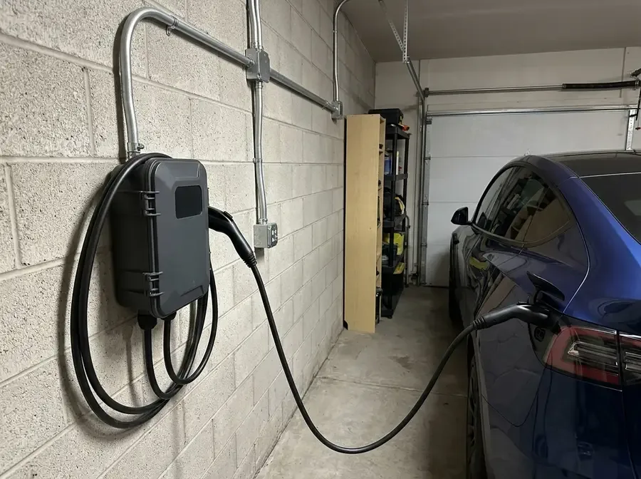 A wall-mounted Level 2 EV charger installed in a garage, its cable plugged into a parked electric car, conduit running neatly from the charger into the wall